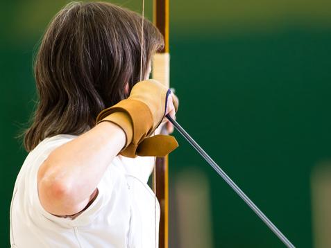 Eine Person in weißem Oberteil und schwarzem Rock zielt mit einem Bogen auf eine Zielscheibe in einer Sporthalle.
