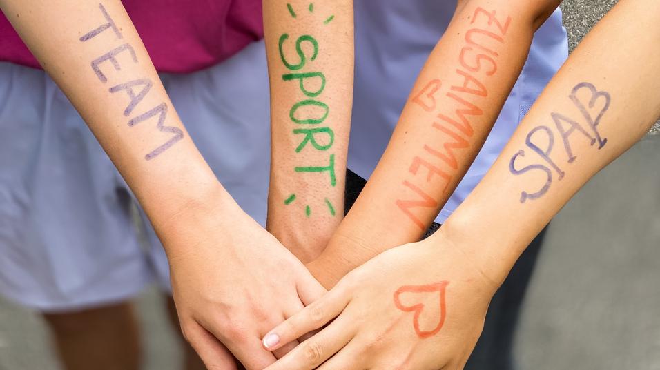 Vier Hände mit bunten Schriftzügen "TEAM", "SPORT", "ZUSAMMEN" und "SPASS" liegen übereinander auf einem grauen Untergrund.